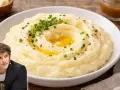 Un hombre sonríe junto a un plato de cremoso puré de patatas, adornado con hierbas frescas y un chorrito de mantequilla derretida.