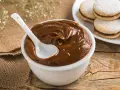 Un tazón blanco lleno de cremoso dulce de leche, con una cuchara blanca sumergida. Al fondo, alfajores espolvoreados con azúcar glas.