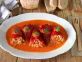 Cinco pimientos rellenos, cubiertos con salsa y perejil, servidos en un plato blanco con pan y cubiertos de madera.