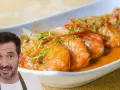 Un hombre sonriente presenta un plato de gambas al ajillo, con gambas jugosas en una salsa naranja, adornadas con finas tiras de chile rojo y hierbas verdes.