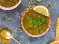 Un tazón de lentejas al curry, adornado con perejil fresco y una rodaja de limón. Una cuchara dorada con cúrcuma completa la escena.
