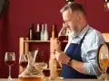 Un experimentado catador de vinos, con barba canosa, huele un cáliz de vino tinto. En la mesa, copas y una jarra con vino.