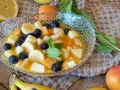 Deliciosa macedonia de frutas con plátano, arándanos, melocotón y manzana en un cuenco de cristal, decorada con menta.