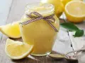 Tarro de lemon curd casero, adornado con un lazo a cuadros, junto a limones frescos y hojas verdes sobre una mesa de madera.