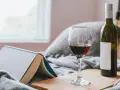 Copa de vino tinto y botella, junto a un libro abierto, sobre una manta gris. Un momento de relax con vino en España.
