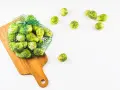 Una bolsa de malla verde llena de coles de Bruselas frescas descansa sobre una tabla de cortar de madera. Algunas coles de Bruselas sueltas se esparcen alrededor.