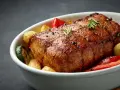 Chuleta de ternera al horno, jugosa y dorada, acompañada de patatas, pimientos rojos y calabacín en una fuente blanca.