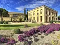 Finca El Caño, un edificio amarillo con arcos y balcones, rodeado de jardines verdes, arbustos y flores moradas bajo un cielo azul con nubes.