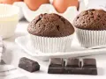 Dos magdalenas de chocolate recién horneadas, listas para disfrutar. Huevos y papelillos de repostería al fondo.