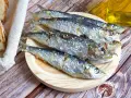 Cinco sardinas al horno, saladas y doradas, reposan en un plato de madera junto a pan rústico y una botella de aceite.