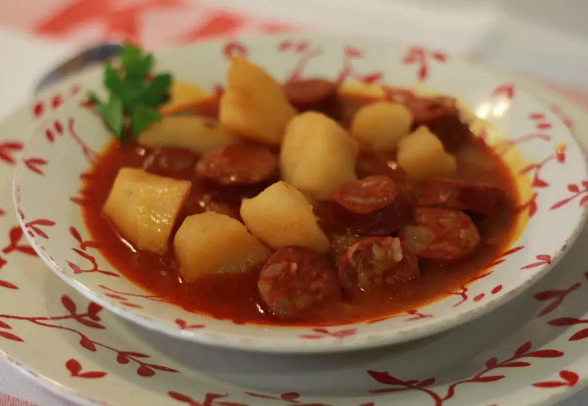 Un plato humeante de patatas a la riojana, con trozos de chorizo y patata en una salsa rojiza, adornado con una ramita de perejil.
