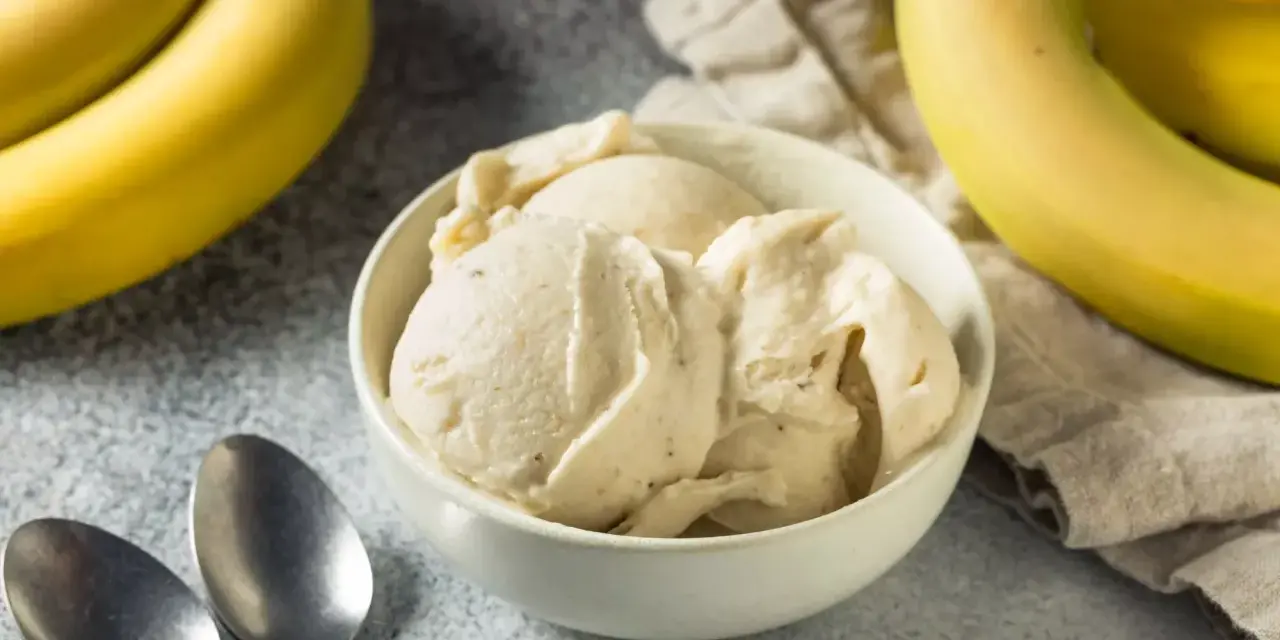 Un cremoso helado de plátano en un tazón blanco, con plátanos amarillos frescos al lado. ¡Perfecto para un postre refrescante!