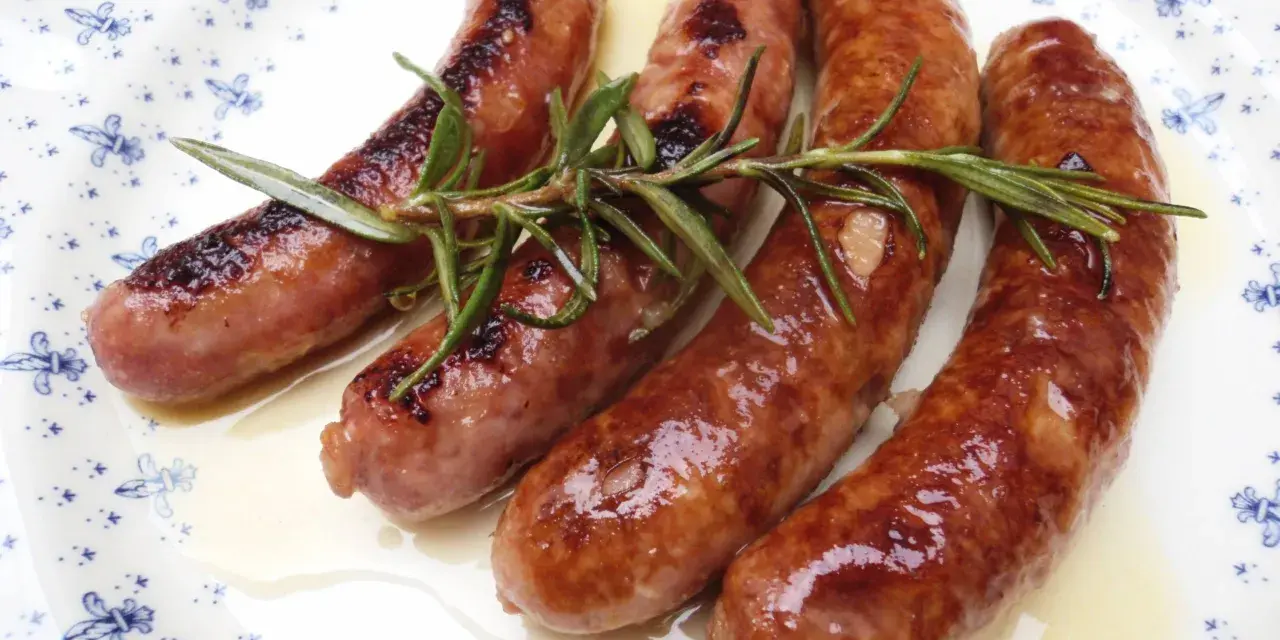 Salchichas al vino Monsieur Cuisine, doradas y jugosas, con romero fresco sobre un plato blanco con motivos azules.