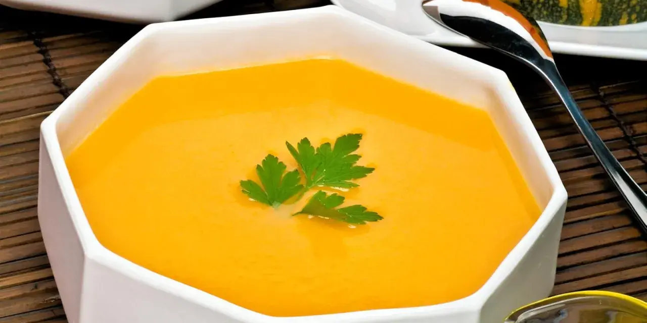 Un cremoso y vibrante **pure de calabaza** en un cuenco blanco, decorado con perejil fresco. Al fondo, un trozo de calabaza y una cuchara.