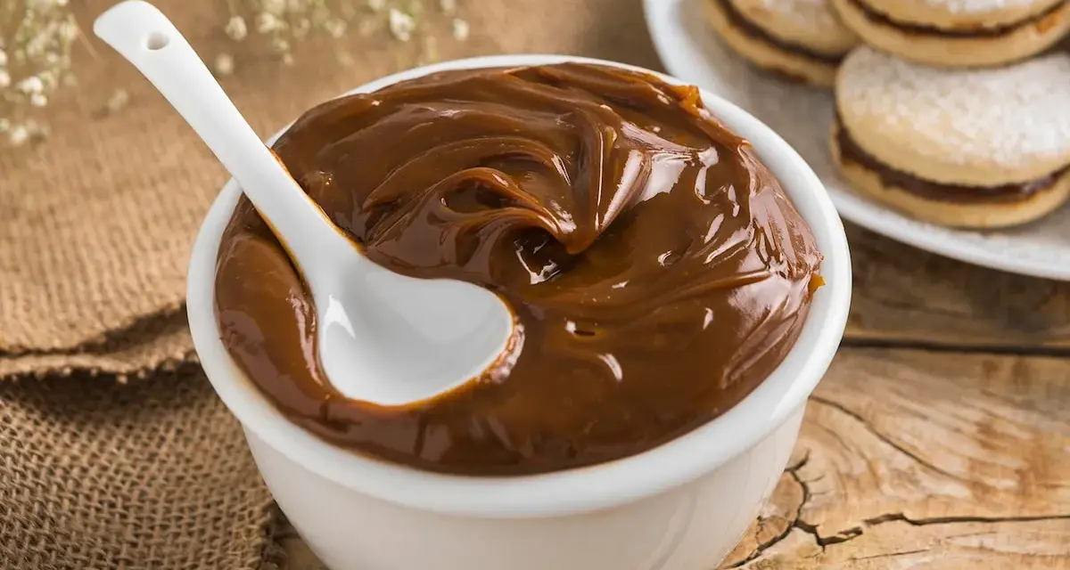 Un tazón blanco lleno de cremoso dulce de leche, con una cuchara blanca sumergida. Al fondo, alfajores espolvoreados con azúcar glas.
