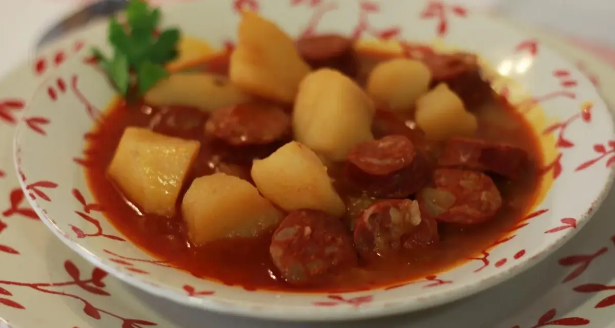 Un plato humeante de patatas a la riojana, con trozos de chorizo y patata en una salsa rojiza, adornado con una ramita de perejil.
