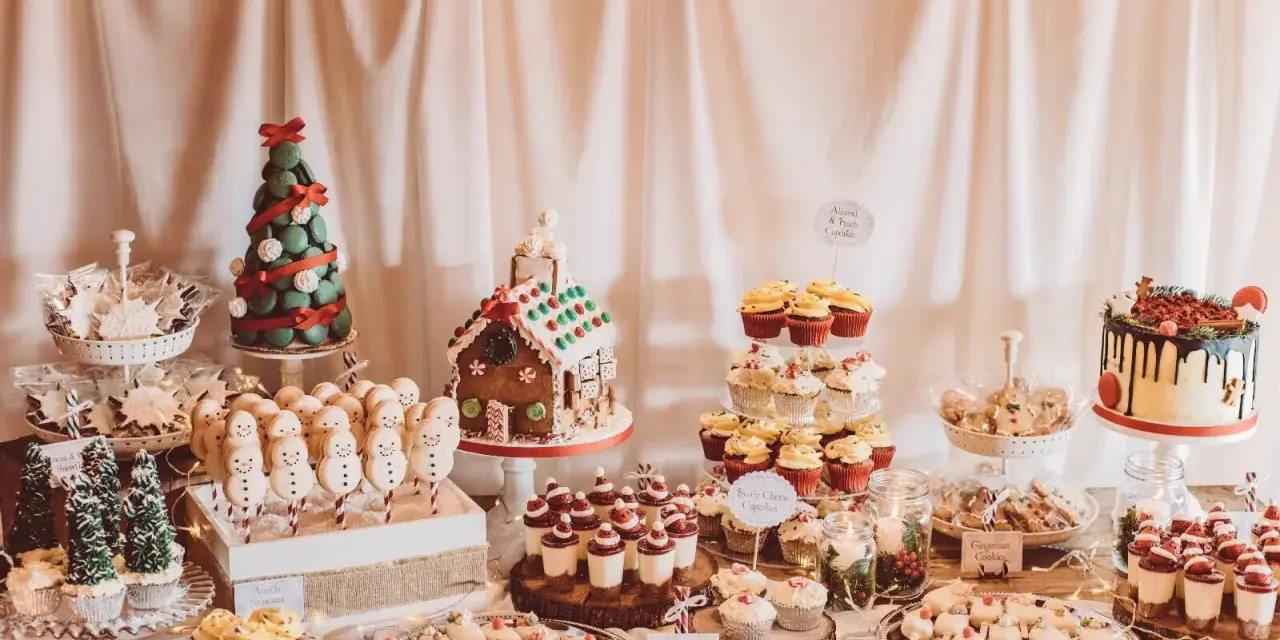Mesa llena de postres navideños: casa de jengibre, árbol de macarons, muñecos de nieve, cupcakes y galletas.