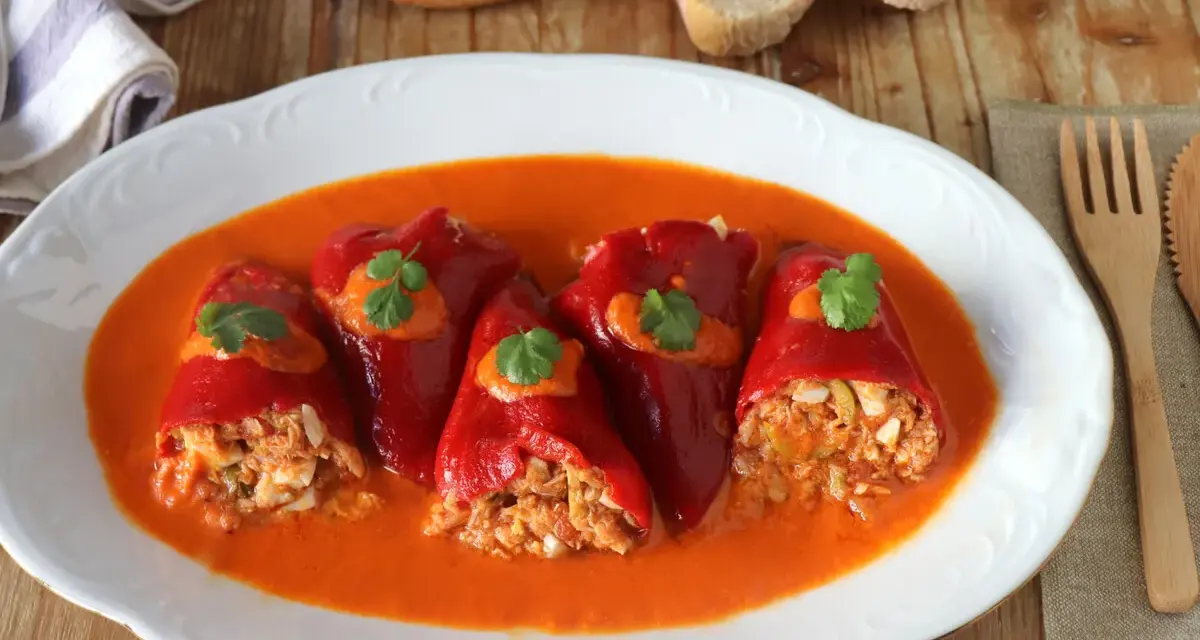 Cinco pimientos rellenos, cubiertos con salsa y perejil, servidos en un plato blanco con pan y cubiertos de madera.