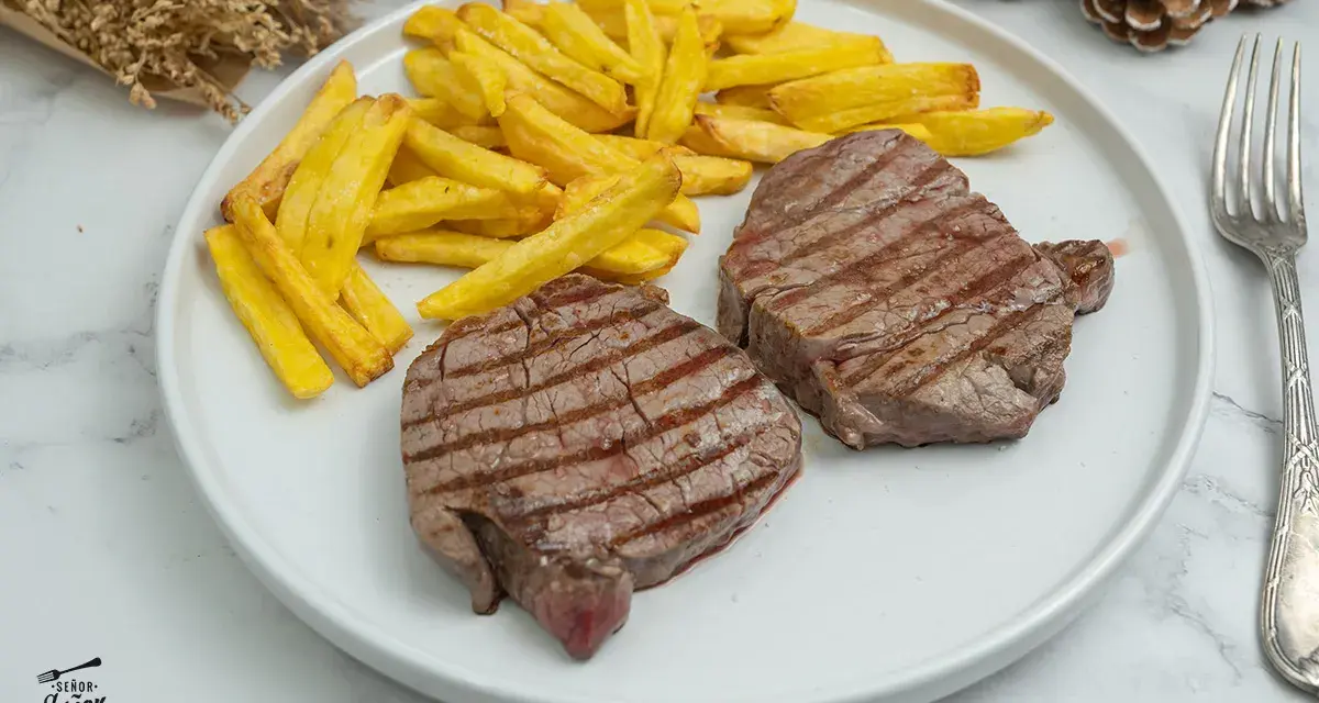 Dos jugosos solomillos de ternera a la plancha, con marcas de parrilla, acompañados de patatas fritas doradas.