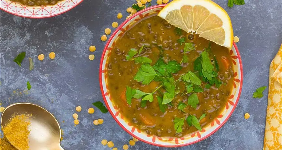Un tazón de lentejas al curry, adornado con perejil fresco y una rodaja de limón. Una cuchara dorada con cúrcuma completa la escena.