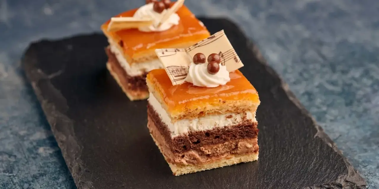 Deliciosa tarta San Marcos en porciones, con capas de bizcocho, crema y cobertura anaranjada, decorada con nata y notas musicales de chocolate.