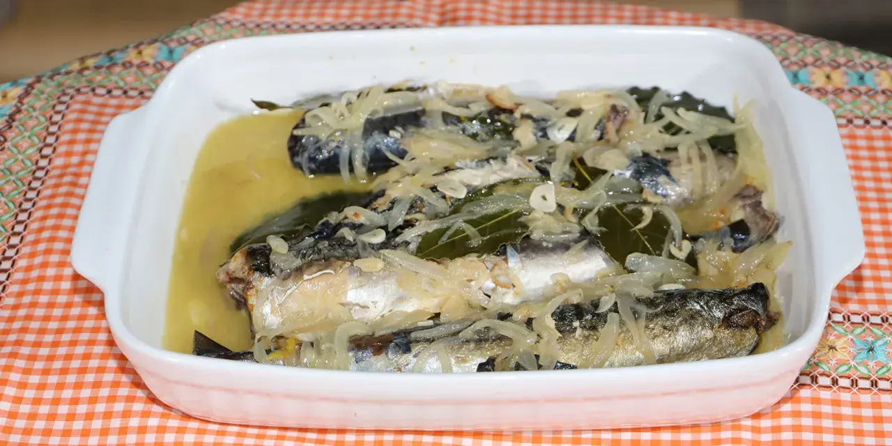 Caballa en escabeche servida en una fuente blanca, con cebolla, laurel y un líquido dorado.