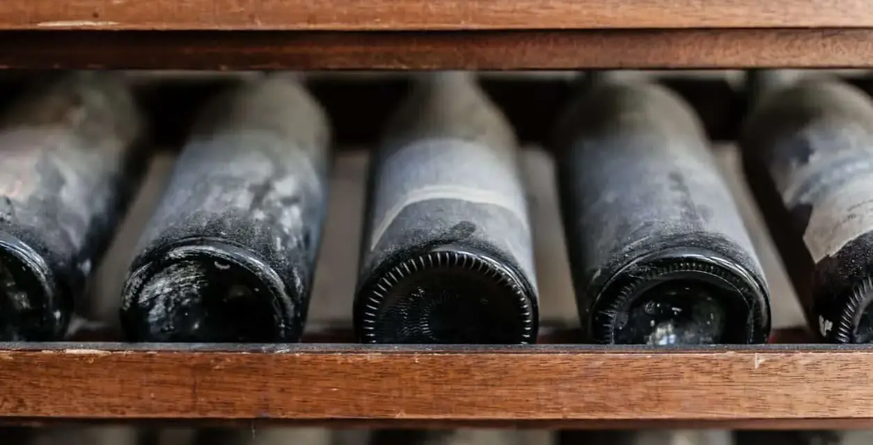 Botellas de vino añejo en estantería de madera, cubiertas de polvo y con el sedimento del vino visible.