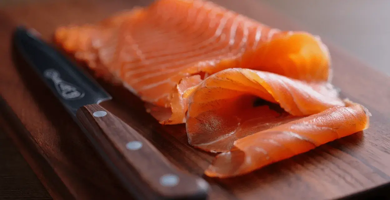 Finísimas lonchas de salmón marinado, listas para disfrutar, junto a un cuchillo de chef sobre tabla de madera.