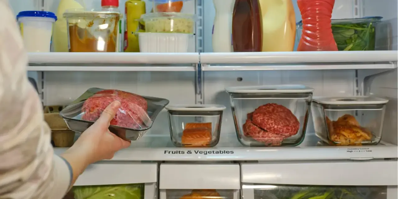 Mano colocando carne picada en la nevera. ¿Cuánto dura la carne en la nevera? También hay salmón, pollo y verduras.
