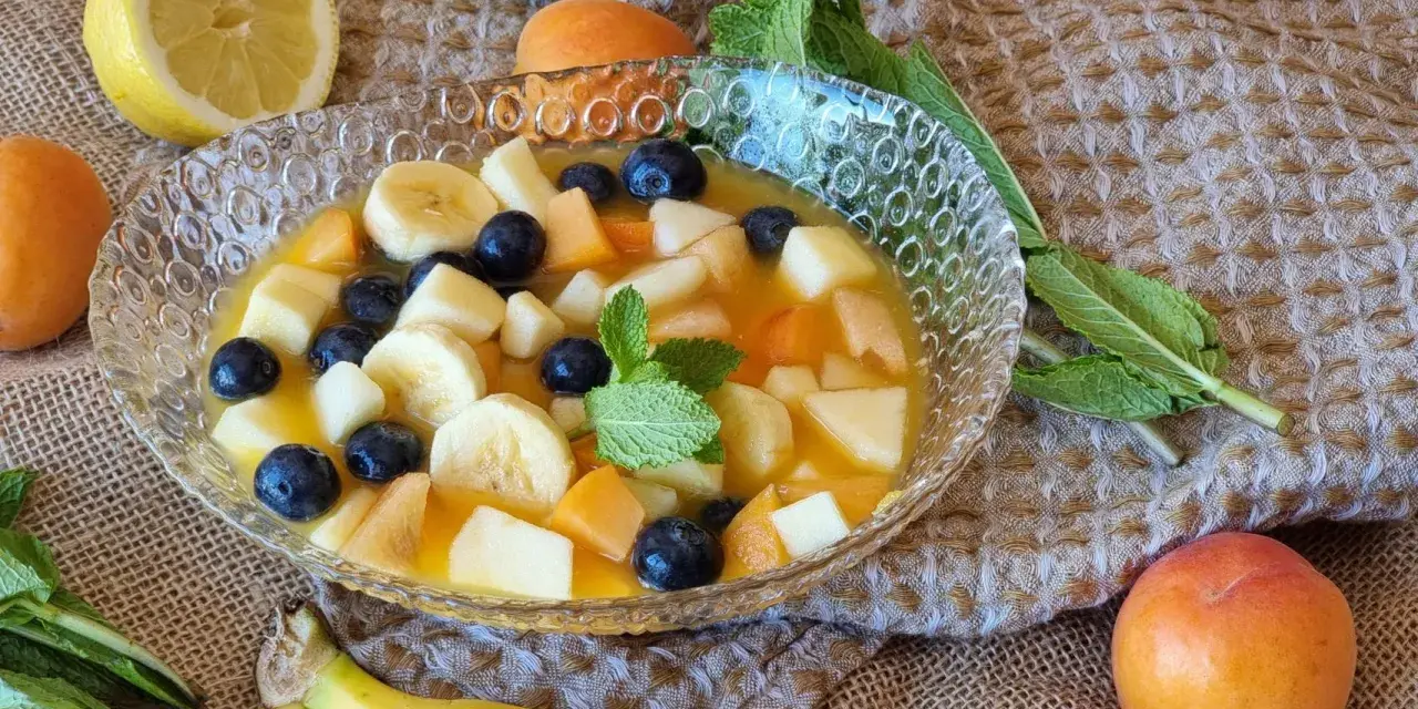 Deliciosa macedonia de frutas con plátano, arándanos, melocotón y manzana en un cuenco de cristal, decorada con menta.