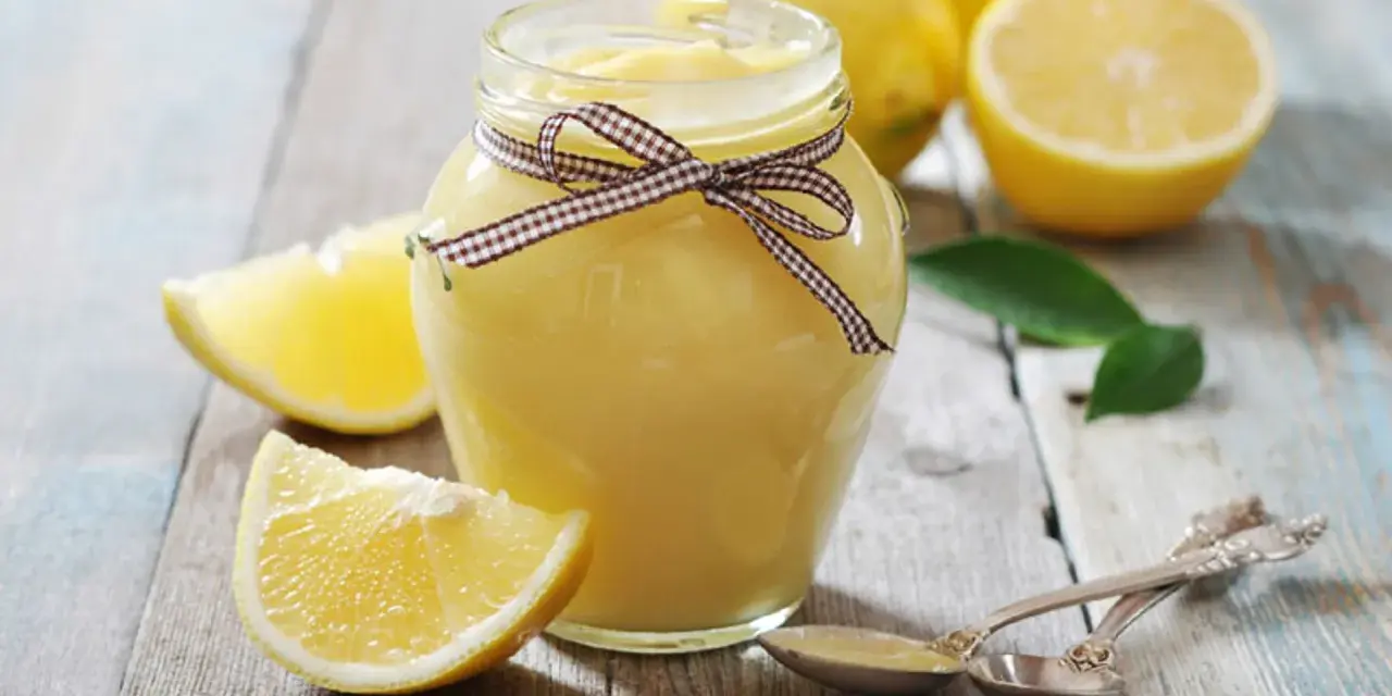 Tarro de lemon curd casero, adornado con un lazo a cuadros, junto a limones frescos y hojas verdes sobre una mesa de madera.