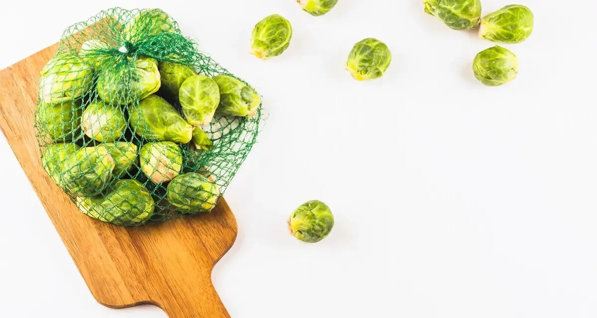 Una bolsa de malla verde llena de coles de Bruselas frescas descansa sobre una tabla de cortar de madera. Algunas coles de Bruselas sueltas se esparcen alrededor.