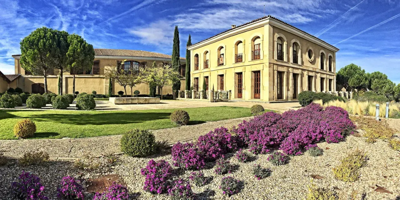 Finca El Caño, un edificio amarillo con arcos y balcones, rodeado de jardines verdes, arbustos y flores moradas bajo un cielo azul con nubes.