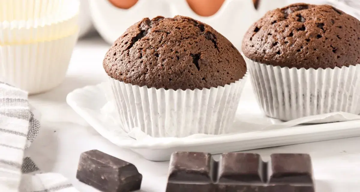 Dos magdalenas de chocolate recién horneadas, listas para disfrutar. Huevos y papelillos de repostería al fondo.