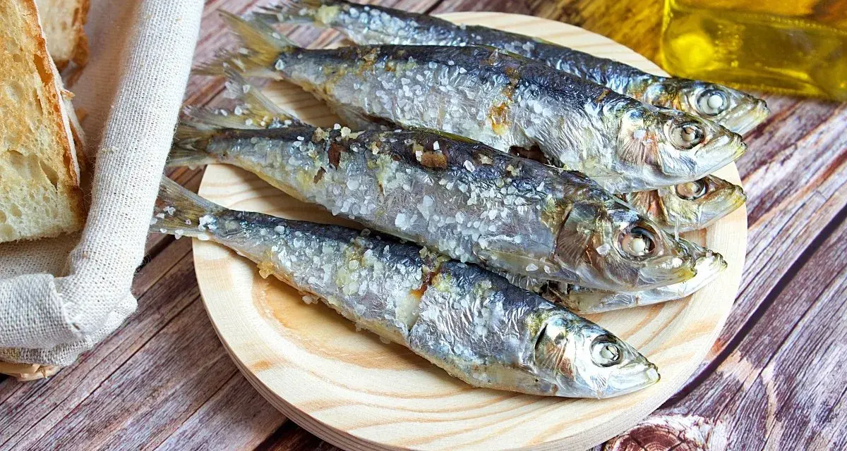 Cinco sardinas al horno, saladas y doradas, reposan en un plato de madera junto a pan rústico y una botella de aceite.