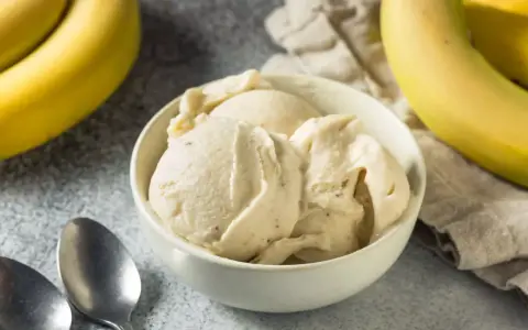 Un cremoso helado de plátano en un tazón blanco, con plátanos amarillos frescos al lado. ¡Perfecto para un postre refrescante!
