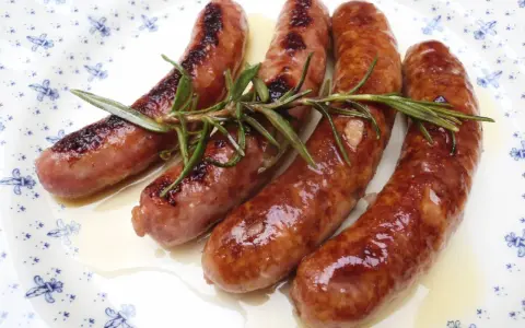 Salchichas al vino Monsieur Cuisine, doradas y jugosas, con romero fresco sobre un plato blanco con motivos azules.