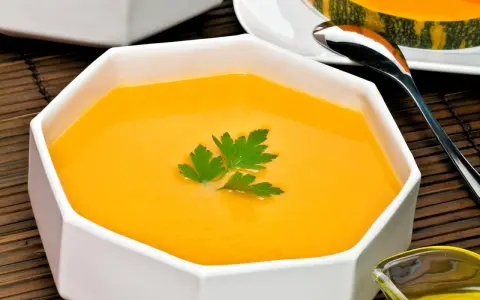Un cremoso y vibrante **pure de calabaza** en un cuenco blanco, decorado con perejil fresco. Al fondo, un trozo de calabaza y una cuchara.