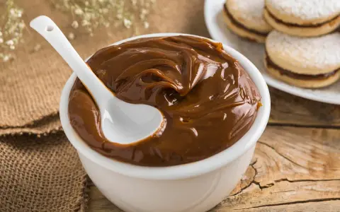 Un tazón blanco lleno de cremoso dulce de leche, con una cuchara blanca sumergida. Al fondo, alfajores espolvoreados con azúcar glas.
