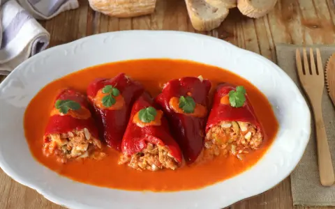 Cinco pimientos rellenos, cubiertos con salsa y perejil, servidos en un plato blanco con pan y cubiertos de madera.