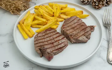 Dos jugosos solomillos de ternera a la plancha, con marcas de parrilla, acompañados de patatas fritas doradas.