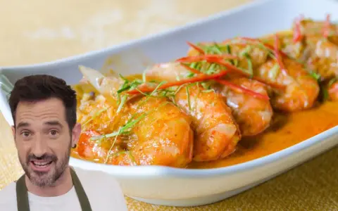 Un hombre sonriente presenta un plato de gambas al ajillo, con gambas jugosas en una salsa naranja, adornadas con finas tiras de chile rojo y hierbas verdes.