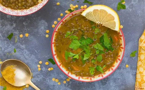 Un tazón de lentejas al curry, adornado con perejil fresco y una rodaja de limón. Una cuchara dorada con cúrcuma completa la escena.