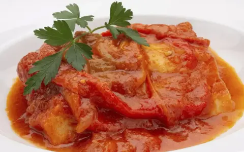 Bacalao a la riojana, un plato tradicional con trozos de pescado blanco cubiertos en una salsa roja de pimientos y tomate, adornado con perejil fresco.