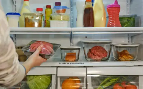 Mano colocando carne picada en la nevera. ¿Cuánto dura la carne en la nevera? También hay salmón, pollo y verduras.