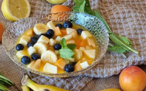Deliciosa macedonia de frutas con plátano, arándanos, melocotón y manzana en un cuenco de cristal, decorada con menta.