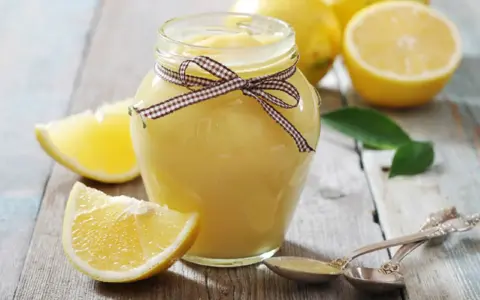 Tarro de lemon curd casero, adornado con un lazo a cuadros, junto a limones frescos y hojas verdes sobre una mesa de madera.