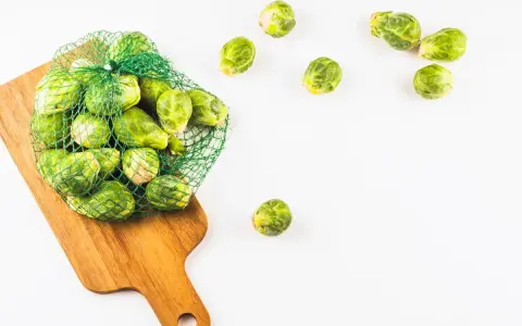 Una bolsa de malla verde llena de coles de Bruselas frescas descansa sobre una tabla de cortar de madera. Algunas coles de Bruselas sueltas se esparcen alrededor.