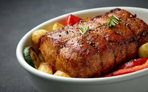 Chuleta de ternera al horno, jugosa y dorada, acompañada de patatas, pimientos rojos y calabacín en una fuente blanca.