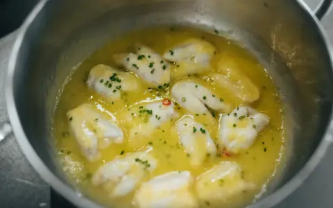 Trozo de bacalao al pil pil burbujeando en una sartén con aceite de oliva, ajo y perejil picado.